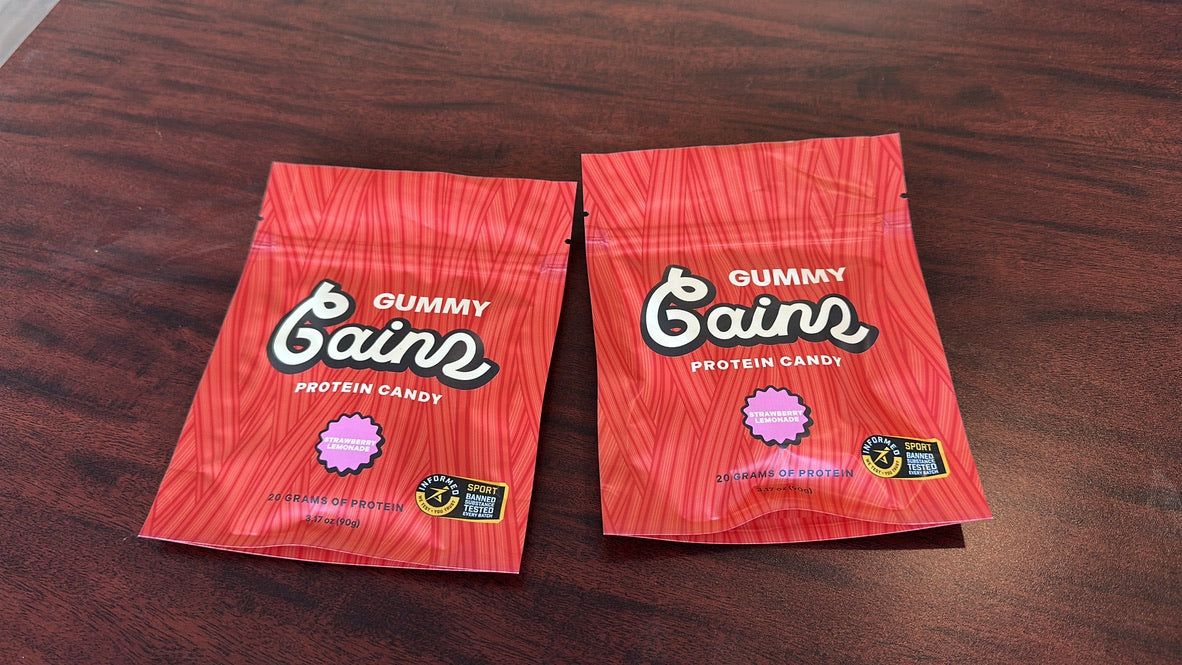 Two Gummy Gainz Strawberry Lemonade protein candy pouches on a wood desk showing 20 grams of protein and the Informed Sport banned-substance tested badge.