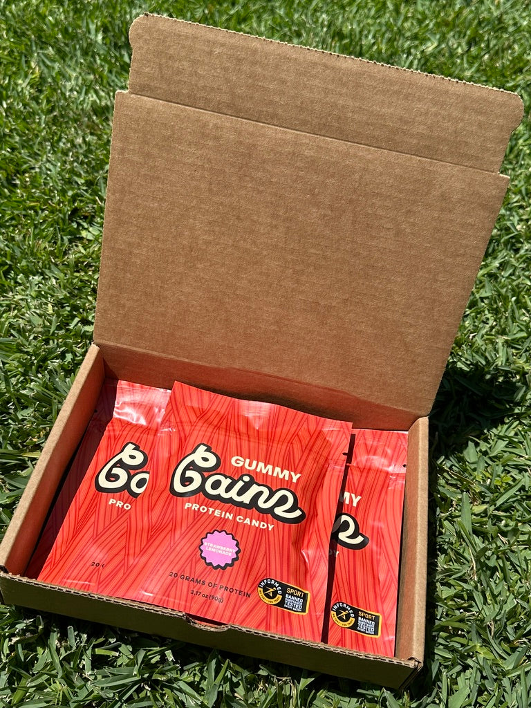 Three Gummy Gainz Strawberry Lemonade protein candy pouches inside an open shipping box on grass — bulk multi-pack unboxing.